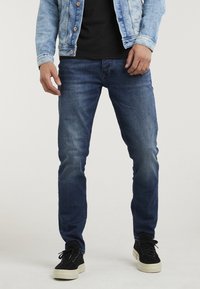 Jeans de mezclilla azul oscuro con un corte ajustado, que presentan un lavado desgastado, estilo de cinco bolsillos, y combinados con zapatillas de ante negras sobre un fondo claro.