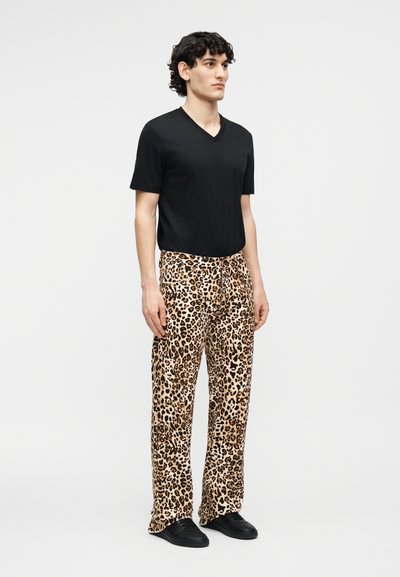 Άνδρας με σγουρά μαλλιά, φορώντας μαύρο T-shirt με λαιμόκοψη σε σχήμα V, παντελόνι με λεοπάρ σχέδιο και μαύρα παπούτσια σε απλό φόντο.