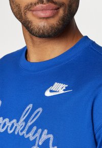 Mėlyna sportinė maikutė su išsiuvinėtu tekstu "Brooklyn" ir balta "Nike" logotipu. Apvali iškirptė ir tekstūruotas audinys. Artimas vaizdas.