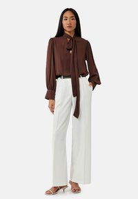 Blusa lunga marrone a maniche lunghe con scollatura annodata, abbinata a pantaloni bianchi a gamba larga e sandali trasparenti con tacco. Tessuto morbido, vestibilità su misura.