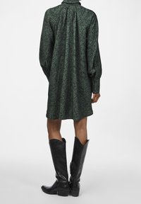 Green patterned dress with a flowing silhouette, pleated back, and long sleeves, paired with tall black leather boots.