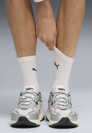 Persoon die witte Puma-sokken aanpast, gedragen met zilveren en roze Puma hardloopschoenen, zichtbaar vanaf de midden-kuit naar beneden tegen een effen achtergrond.