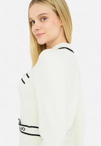 Maglione bianco lavorato a maglia con dettagli e strisce nere, caratterizzato da un colletto rotondo e una vestibilità aderente. Texture liscia, visibili maniche lunghe.