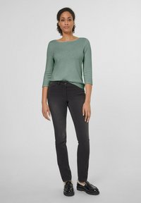 Grüner, langärmeliger Pullover mit weit geschnittenem Ausschnitt, kombiniert mit schwarzen, figurbetonten Jeans und schwarzen Schuhen, getragen von einer Person in einer neutralen Umgebung.