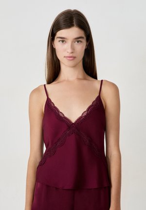 Femme portant un haut sans manches bordeaux avec une bordure en dentelle au niveau de l'encolure et des lignes diagonales sur le devant.