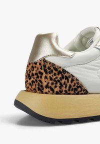 Zapatilla con una parte superior sintética blanca, talón metálico dorado y un panel texturizado de estampado de leopardo. Suela de goma negra gruesa con acentos amarillos.