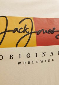 Bílý tričko s velkým černým kurzivním logem "Jack & Jones", nad nímž jsou žluté a červené barevné bloky, a pod ním černý text "ORIGINAL WORLDWIDE".