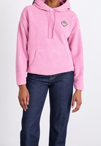 Persoon in een roze fleece hoodie met een voorzak en donkere spijkerbroek tegen een plain witte achtergrond.