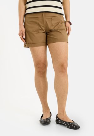 Mujer con pantalones cortos marrones remangados, bailarinas negras con tachuelas, camiseta de rayas y pulseras, de pie frente a un fondo blanco liso.