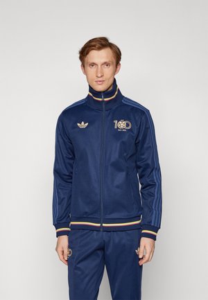 Giacca a zip navy in tessuto felpato con logo dorato, tre linee sulle maniche e colletto e polsini a righe multicolori.