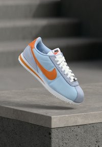 Nike tenisice sa svijetloplavim i sivim gornjim dijelom, narančastim nagliscima, bijelim vezicama i teksturiranom gumenom potplatom. Sportski dizajn s logotipom.