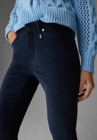 Marinblå, figursydda byxor i mjukt bomullsmaterial. Har dragkedja fram, knappstängning och framfickor. Texturerad stickad tröja synlig.