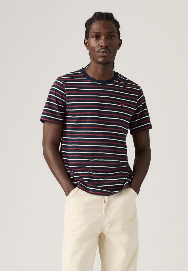 Mann mit geflochtenem Haar, der ein dunkelblaues, rot-weiß gestreiftes T-Shirt und hellbeige Hosen trägt, steht mit den Händen in den Taschen.