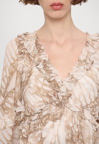 Femme portant un chemisier beige et blanc à motifs avec des détails à volants le long de l'encolure en V et de la zone de la poitrine.