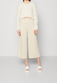Romige, cropped textuurcardigan met knoopsluiting, gecombineerd met beige, wijde broekspijpen. Witte plateausandalen maken de outfit compleet.