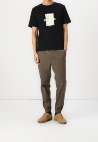 Mężczyzna w czarnej koszulce z grafiką, brązowych spodniach dresowych i beżowych butach sportowych stoi na tle białej ściany.