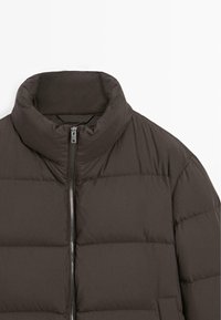 Puchowa kurtka w ciemnym brązie, z wysokim kołnierzem i przednim zamkiem błyskawicznym. Gładka faktura materiału z widocznym wzorem pikowania i minimalnym przeszyciem.