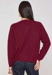 Maglione bordeaux con una vestibilità comoda, maniche raglan e polsini a coste, abbinato a jeans blu scuro. Texture soffice e design semplice.