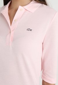 Polo tričko v světle růžové barvě s texturovaným materiálem, s klasickým límcem, dvěma knoflíky a vyšitým logem krokodýla na hrudi.