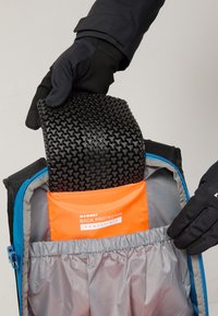 Zwarte flexibele rugbeschermer met een gestructureerd oppervlak, gehouden boven een oranje compatibele hoes in een grijze rugzakcompartiment met een blauwe rits.