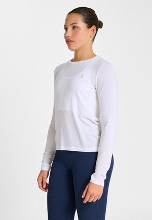 Kvinna står, klädd i en vit långärmad träningsshirt och marinblå träningsleggings, tittar något åt vänster mot en enkel bakgrund.