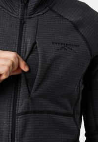 Eine Hand öffnet eine mit Reißverschluss versehene Brusttasche an einer dunkelgrauen strukturierten Jacke mit "SWEDMOUNT" und einem Berglogo auf der Brust.