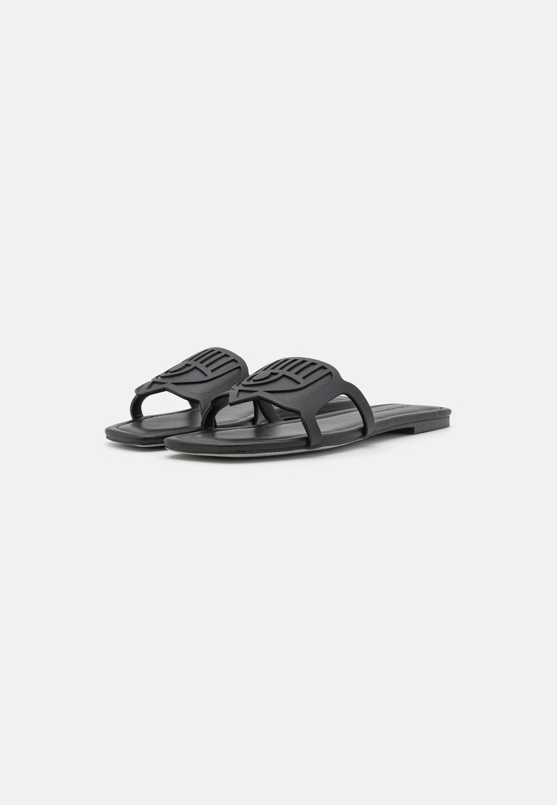 Ein Paar schwarze flache Sandalen mit einem breiten Riemen, der ein stilisiertes Augendesign aufweist, vor einem weißen Hintergrund.