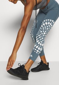 Leggings teal a vita alta con un motivo di pois irregolari bianchi, abbinati a scarpe sportive nere. Il tessuto sembra elastico e aderente.
