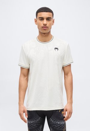 Νεαρός άνδρας που φοράει λευκό κοντομάνικο πουκάμισο με λογότυπο μαύρου φιδιού στο στήθος και μαύρο κολάν με σχέδια.