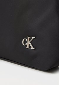 Juodas nailono krepšys su sidabriniu metaliniu cK logotipu. Paviršius lygus, matinio finišo, ir jis turi struktūrizuotą formą.