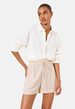 Vrouw in een wit overhemd met knopen en opgerolde mouwen en beige short met trekkoord, staand voor een effen achtergrond en zijwaarts kijkend.