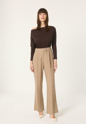 Femme debout portant un haut marron foncé épaules dénudées, un pantalon beige taille haute à jambes larges, et des talons aiguilles beiges sur fond blanc.
