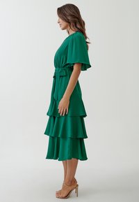 Green tiered dress made of lightweight fabric, featuring a belted waist, short sleeves, and layered ruffles, complemented by nude high-heeled shoes.