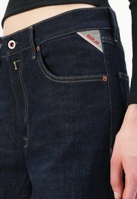 Donkerblauwe denim jeans met contrasterende gele stiksels, metalen hardware, voorzakken en een driehoekige rode en grijze logopatch.