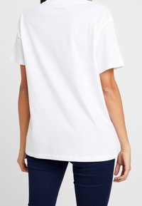 Camiseta de algodón blanca con corte relajado, mangas cortas y cuello redondo. Pequeño logo de la marca impreso en la parte posterior cerca del cuello.