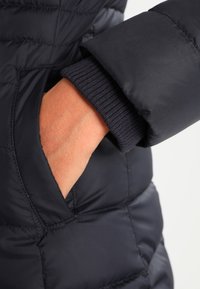 Navy puffer jacket featuring a ribbed cuff, with a hand placed in a side pocket. The fabric is smooth with a quilted texture.