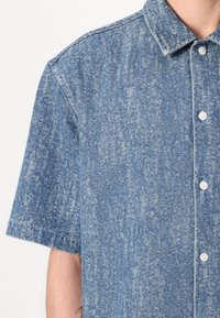 Camicia a maniche corte con bottoni in tessuto di denim a motivo blu. Caratterizzata da un colletto classico e bottoni bianchi, con una finitura texturizzata e leggermente sbiadita.