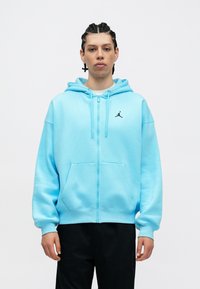 Ανοιχτό μπλε φλιτζάνι φόρμα με κοντά μανίκια και καπιτονέ ύφασμα, διαθέτει τσέπη καγκουρό και μικρό μαύρο λογότυπο Jordan στην αριστερή πλευρά του στήθους.