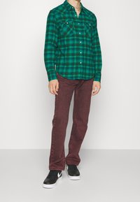 Persona vestida con una camisa de cuadros verdes, jeans burdeos, zapatillas Nike en blanco y negro, y un collar de cadena, de pie contra una pared simple.