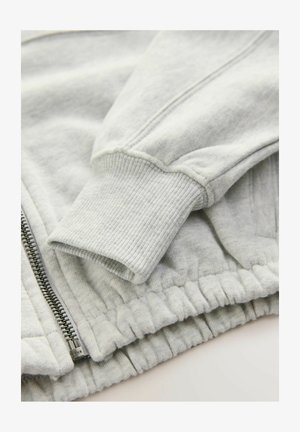 Hanorac gri deschis cu manșete și talie ribbate, având un fermoar complet pe față, realizat dintr-un material moale și texturat.