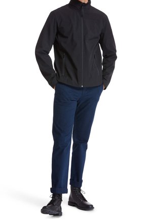 Black softshell jacket with a high collar, zippered pockets, and a fitted cut, paired with navy blue pants and black lace-up boots.