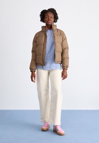 Giacca a vento marrone con colletto alto, t-shirt a righe blu e bianche, pantaloni a palazzo crema e sneakers rosa con dettagli verdi.