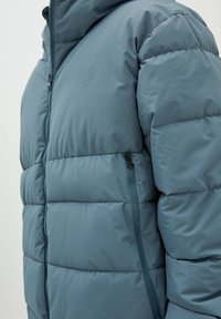 Vadderad blå jacka med dragkedja, med hög krage och en diskret logotyp på bröstet. Slät yta och sidofickor med dragkedja.