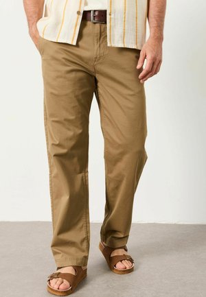 Mann trägt beige Hose, braunen Gürtel, gestreiftes Hemd und braune offene Sandalen, steht auf grauem Boden mit Händen in den Taschen.