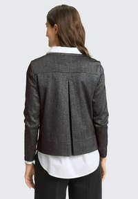 Czarna kurtka o metalicznym połysku z długimi rękawami, detalem rozcięcia z tyłu i dopasowanym fasonem. Noszona na białej koszuli z kołnierzykiem.