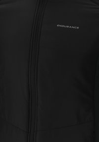 Sort atletisk jakke med frontlynlås, lavet af glat, fleksibelt stof. Har et logo trykt i sølv og et minimalistisk design.