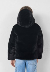 Chaqueta de piel sintética negra con capucha, que presenta una textura suave y un corte holgado. Combinada con unos vaqueros ligeros de corte amplio. Vista trasera.