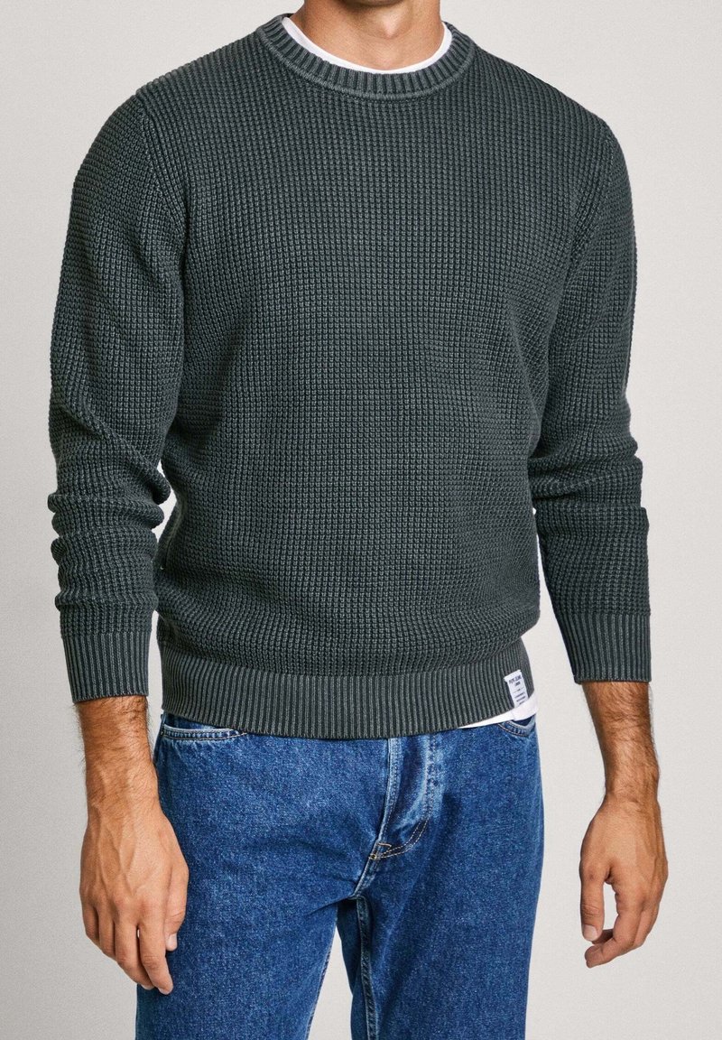 Muž v tmavě zeleném texturovaném pleteném svetru a modrých džínách stojí s rukama volně podél těla proti jednobarevnému pozadí.