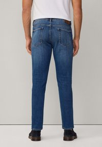 Jeans de mezclilla azul con corte recto, que presentan dos bolsillos traseros y una etiqueta de cuero en la cintura. Botas marrón oscuro visibles en los pies.
