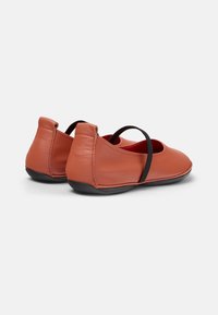 Bruna läderballettskor med ett svart elastiskt band över toppen, vilket visar en enkel, elegant design och en slät, rund form.